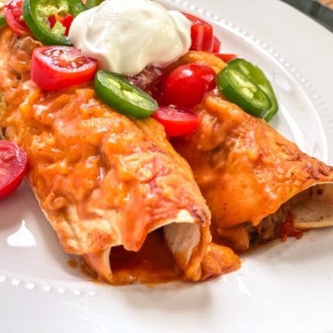 Cheesy enchiladas on a plate with tomatoes, jalapeno, and sour cream.